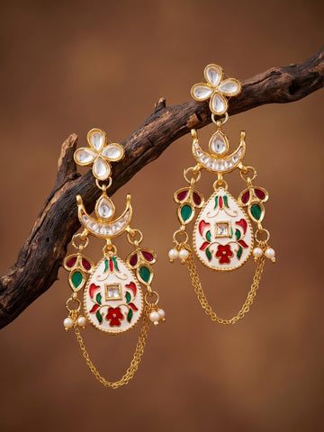 A pair of ornate gold-toned Indian jhumka earrings with white stone floral tops, crescent moon details, and white enamel pendants decorated with red and green floral enameled motifs, hanging from a dark brown branch against a warm brown background.