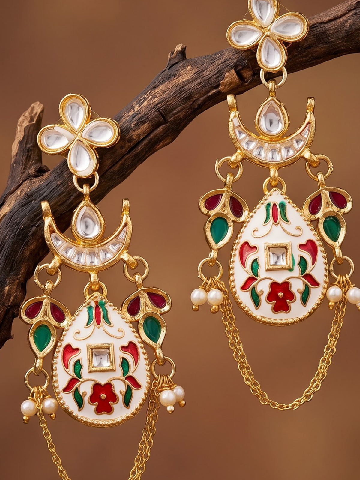 A pair of ornate gold-plated Indian style earrings with white enamel, red and green floral engravings, and small pearl-like beads, hanging from a dark brown branch against a warm brown background.