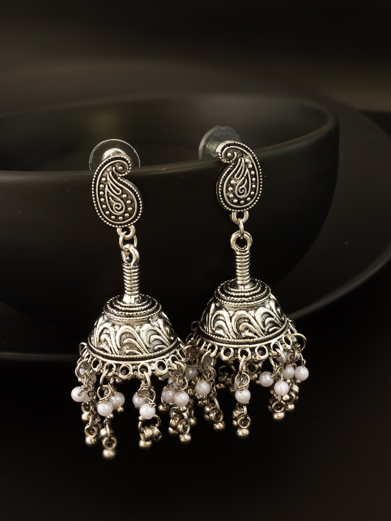 A pair of ornate silver-toned jhumka earrings with paisley-shaped studs, intricate filigree detailing, and dangling pearl-like beads, displayed against a dark background.