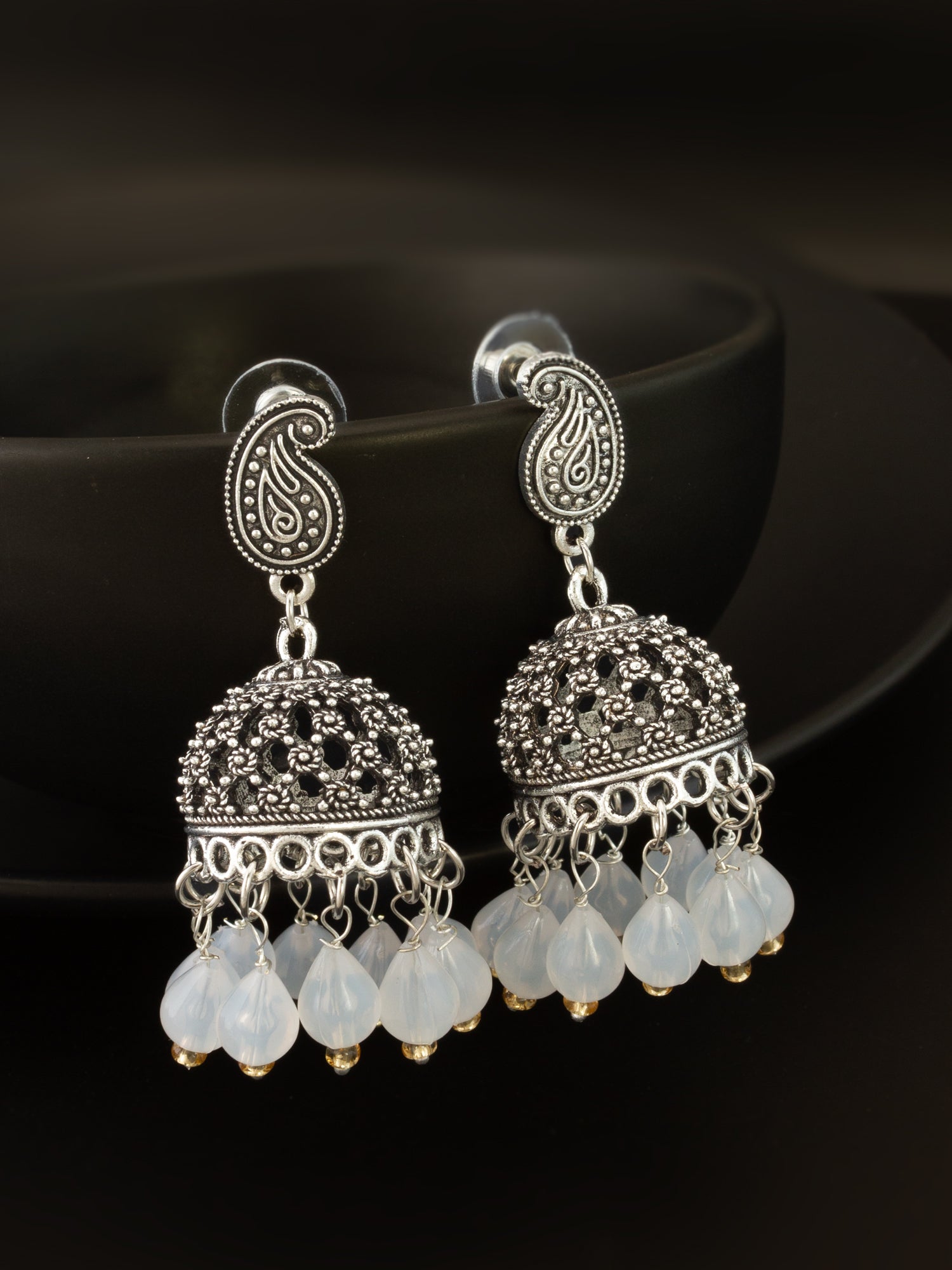 A pair of ornate silver-toned jhumka earrings with intricate paisley and filigree details, featuring dangling translucent white beads, displayed against a dark background.
