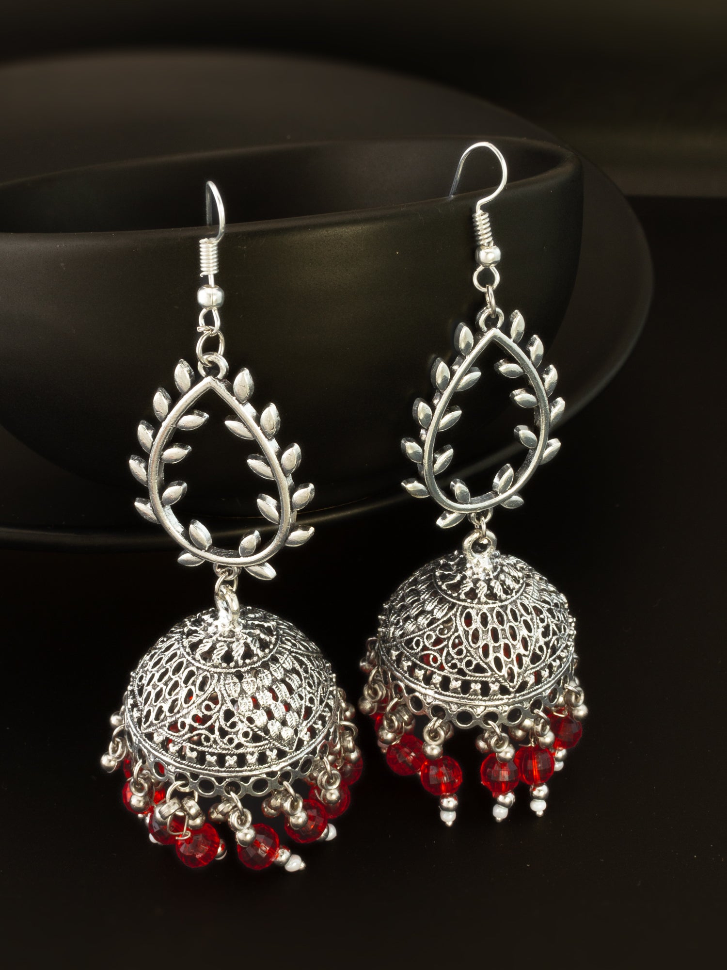 A pair of silver-toned Indian jhumka earrings with red bead accents, displayed against a dark background.