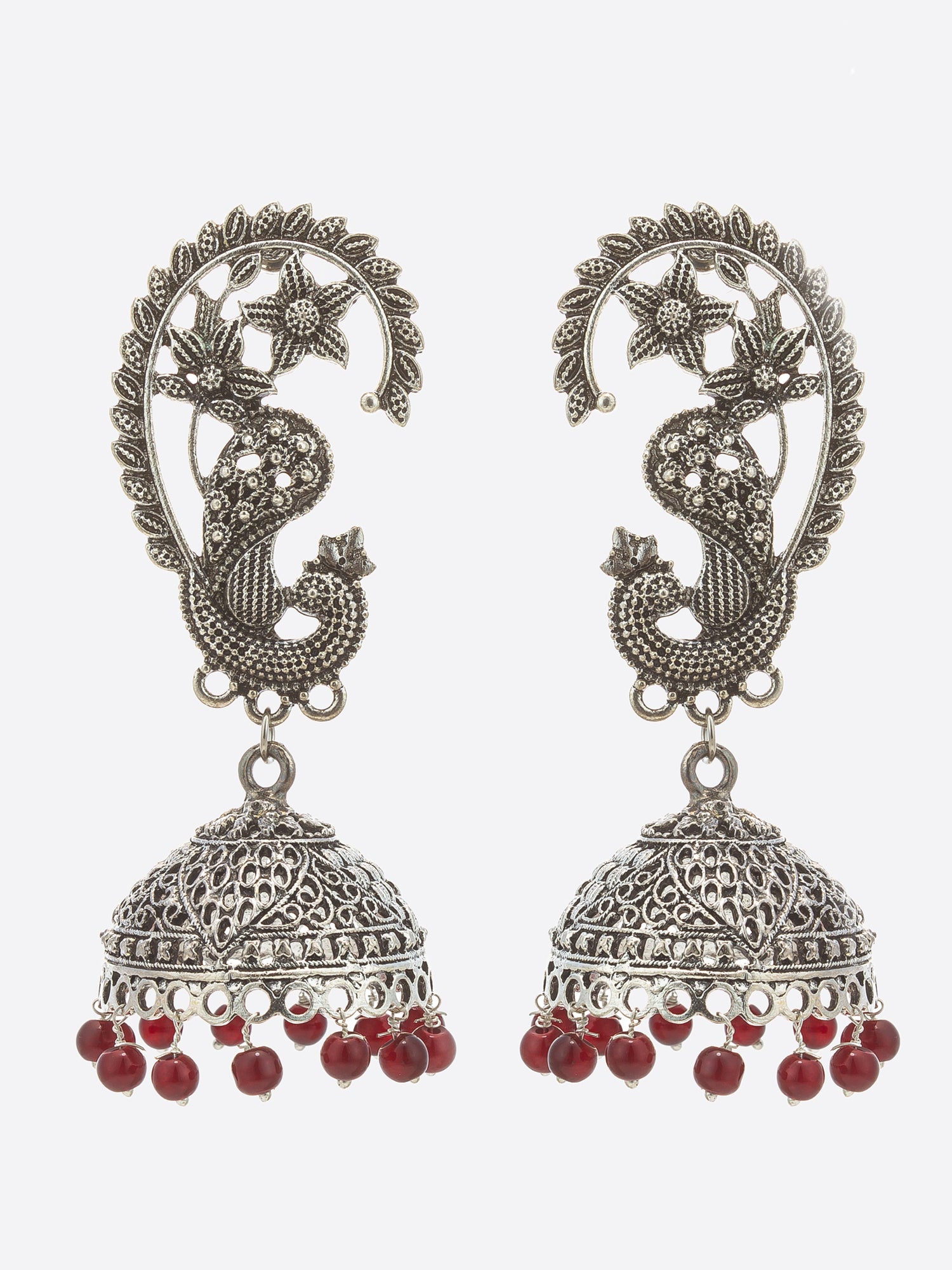A pair of traditional Indian silver-toned jhumka earrings with detailed paisley and peacock motifs, decorated with small clear stones and finished with dangling red beads, shown against a light gray background.