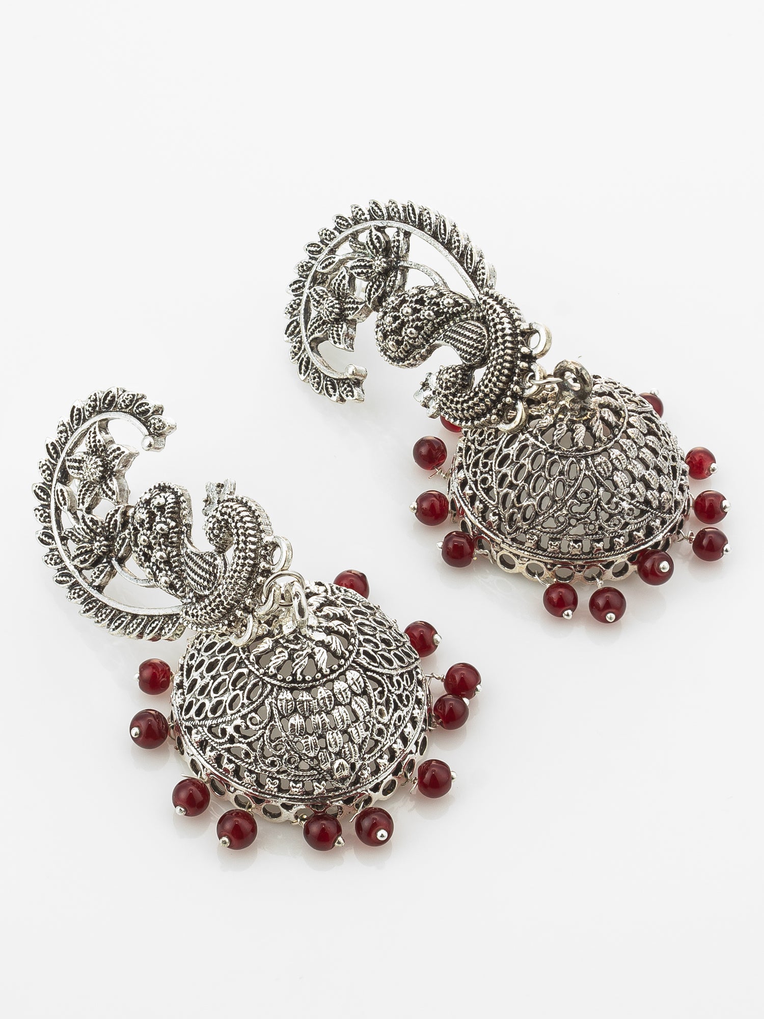 A pair of ornate silver-toned jhumka earrings with detailed filigree work, featuring a peacock motif on the upper hook and small red beads dangling from the bottom, shown on a plain white background.