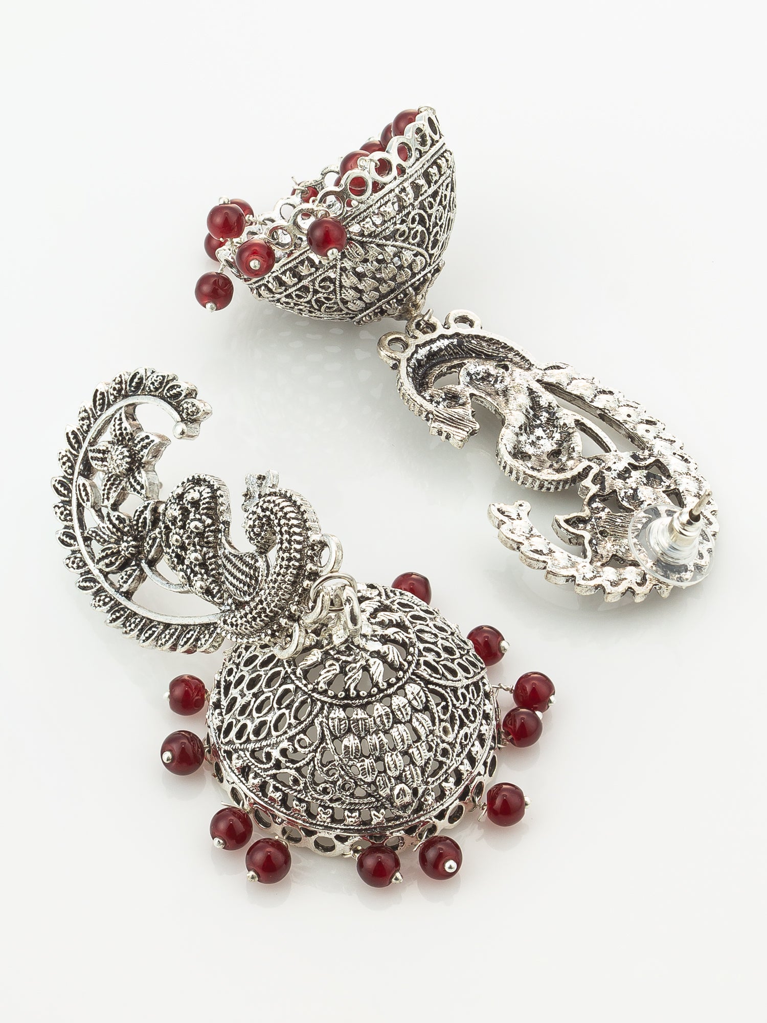 A pair of ornate silver-toned traditional Indian jhumka earrings decorated with intricate filigree work and accented by small red beads, displayed on a clean white background.