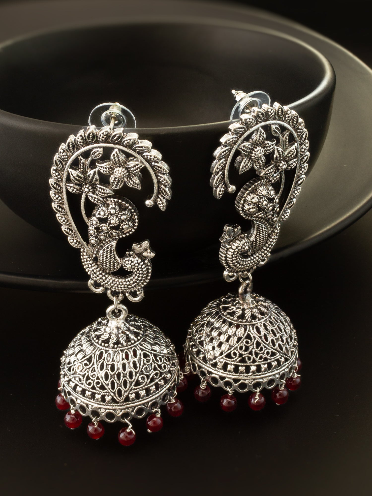 A pair of silver-toned Indian jhumka earrings with detailed filigree work, peacock motifs, and small red bead tassels, displayed against a dark background.