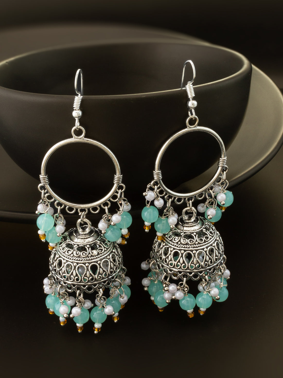 A pair of silver-toned jhumka earrings with intricate filigree detailing, decorated with light blue-green beads and small white pearl-like accents, displayed against a dark background.