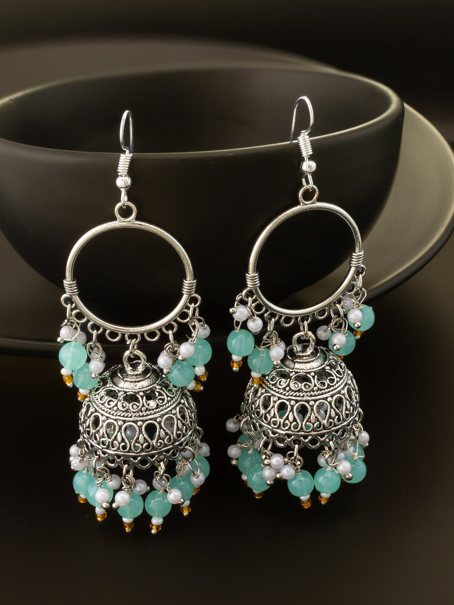 A pair of silver-toned jhumka earrings with intricate filigree detailing, decorated with light blue-green beads and small white pearl-like accents, displayed against a dark background.