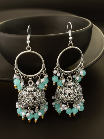 A pair of silver-toned jhumka earrings with intricate filigree detailing, decorated with light blue-green beads and small white pearl-like accents, displayed against a dark background.