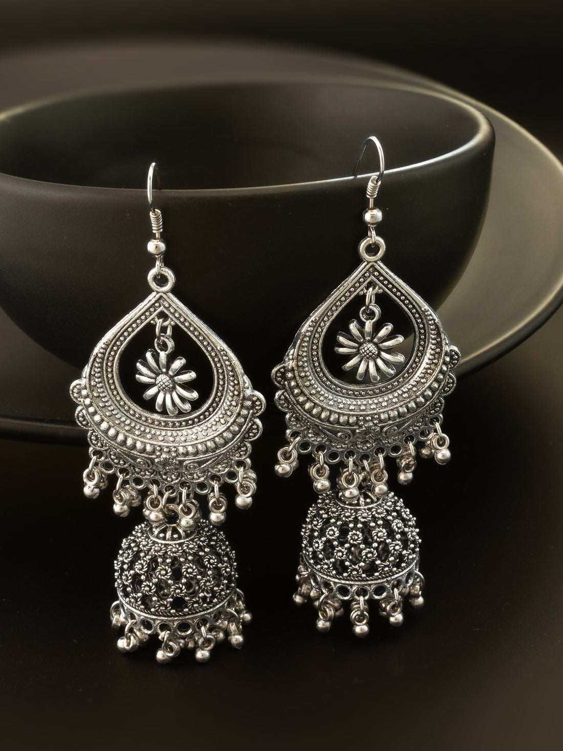 A pair of ornate silver-toned Indian jhumka earrings with intricate filigree work and dangling bell-shaped charms, displayed against a dark background.