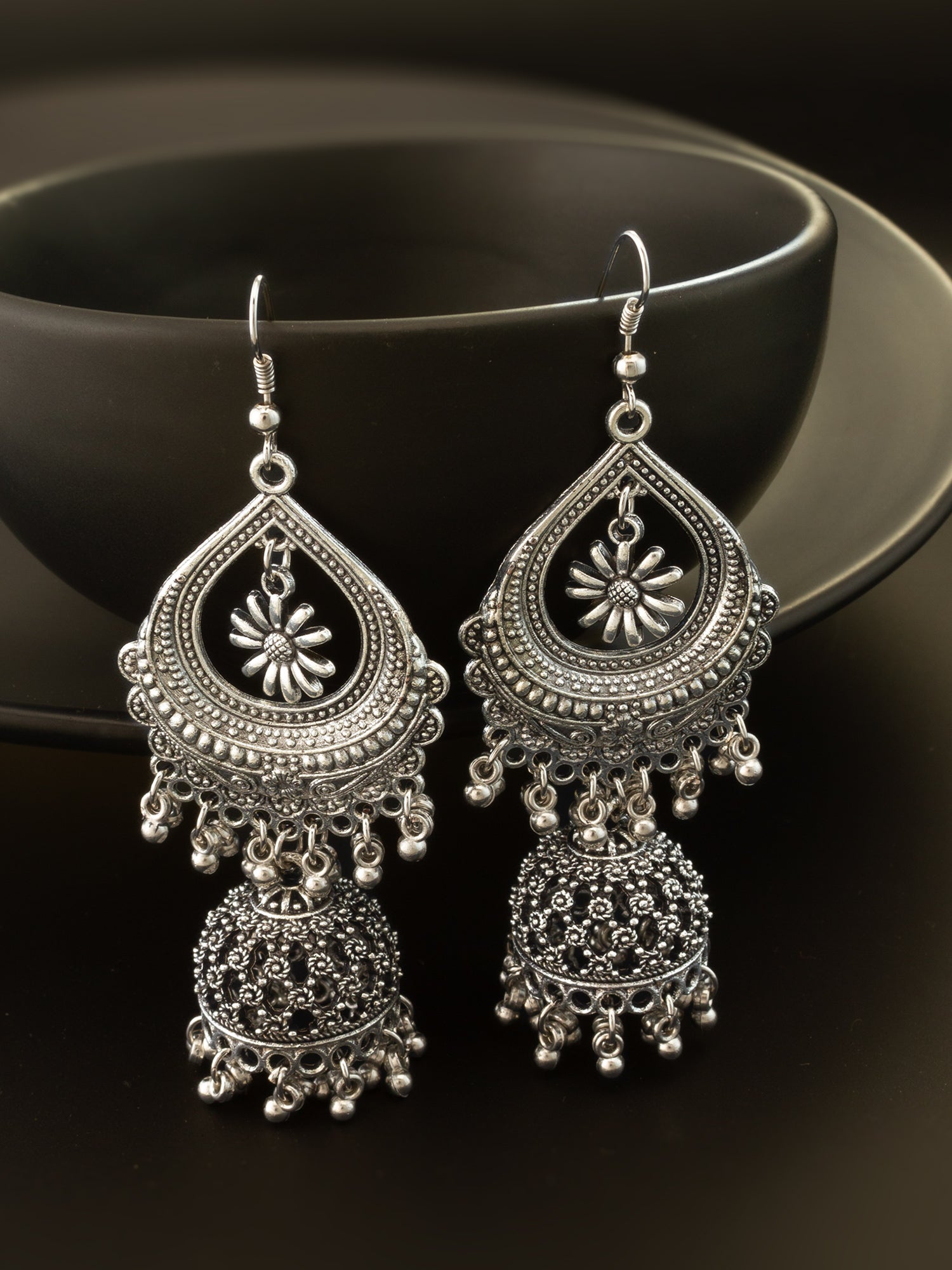 A pair of ornate silver-toned Indian jhumka earrings with intricate filigree work and dangling bell-shaped charms, displayed against a dark background.
