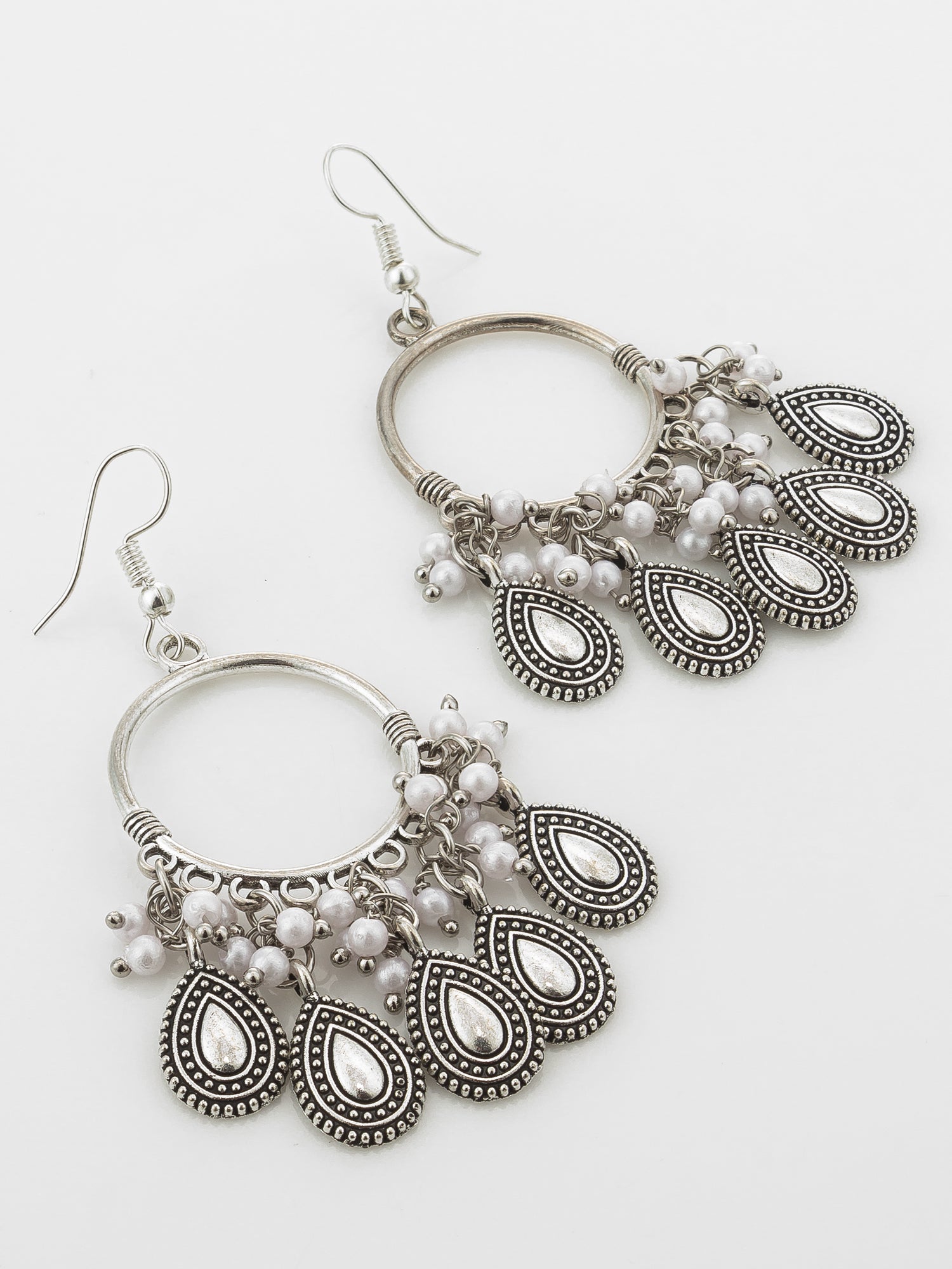 A pair of silver-toned huggie hoop dangle earrings with ornate filigree teardrop charms, small white pearl-like beads, and silver fishhook wires, shown on a plain white background.