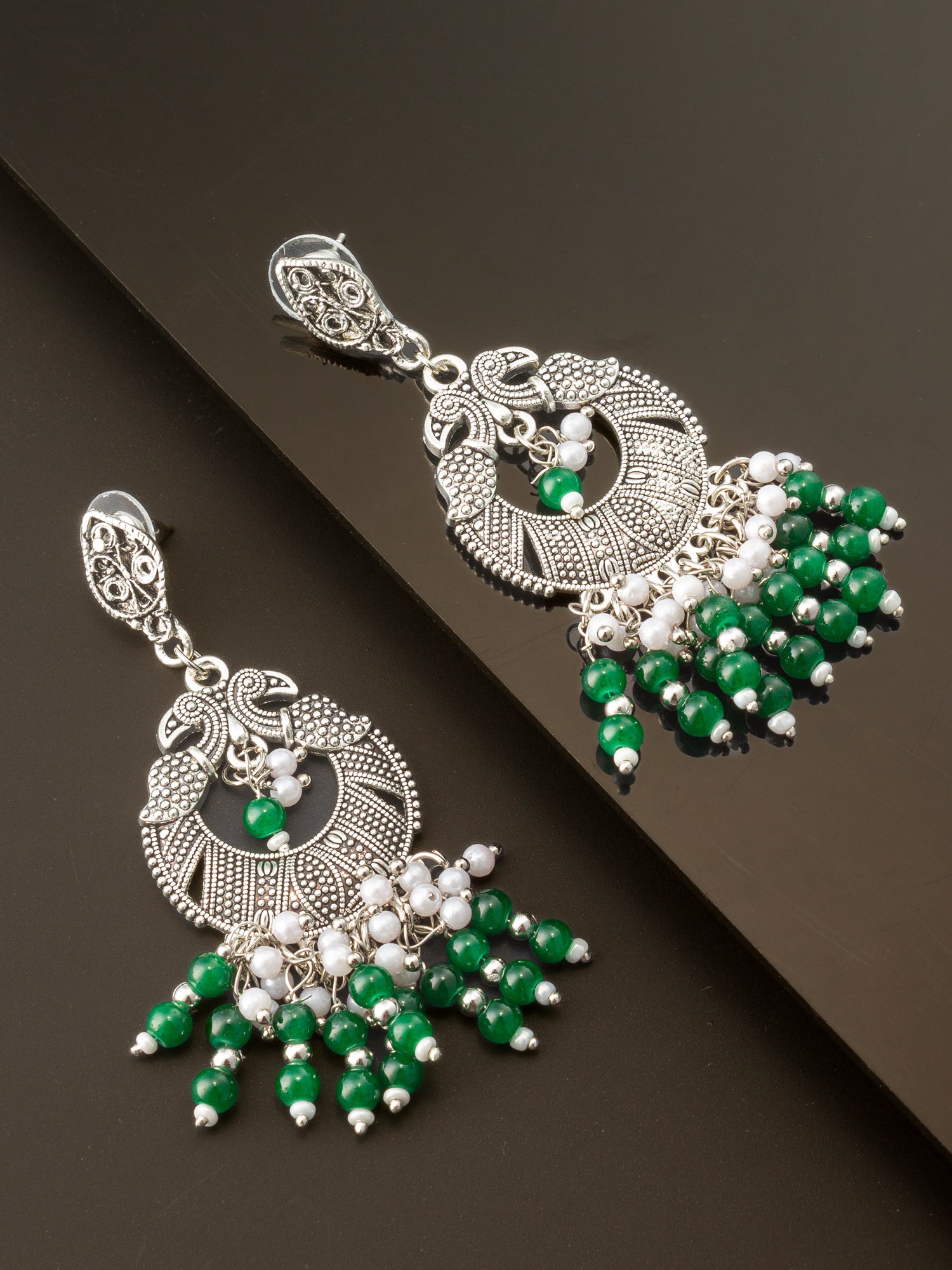 A pair of silver-toned traditional Indian chandbali earrings decorated with green and white beads, displayed on a dark reflective surface.
