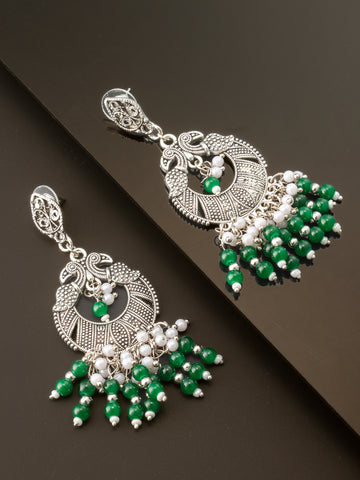 A pair of silver-toned traditional Indian chandbali earrings decorated with green and white beads, displayed on a dark reflective surface.