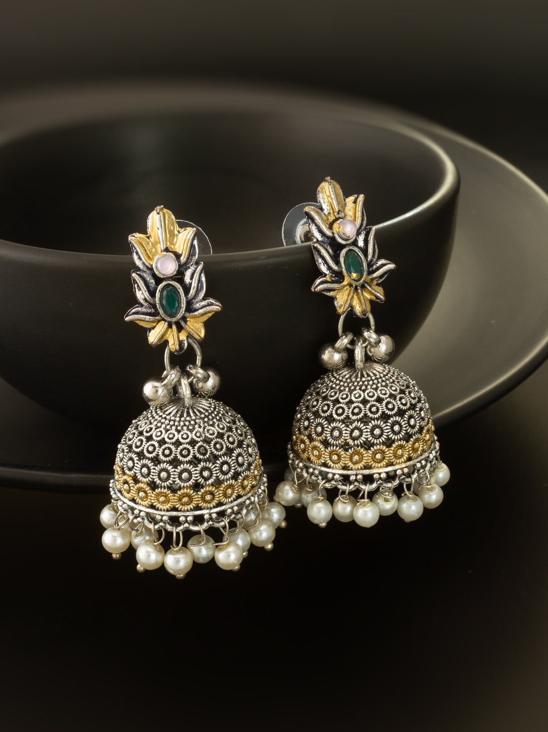 A pair of ornate Indian jhumka earrings with silver filigree bells, gold accents, and small pearl drops, displayed on a black ceramic dish against a dark background.