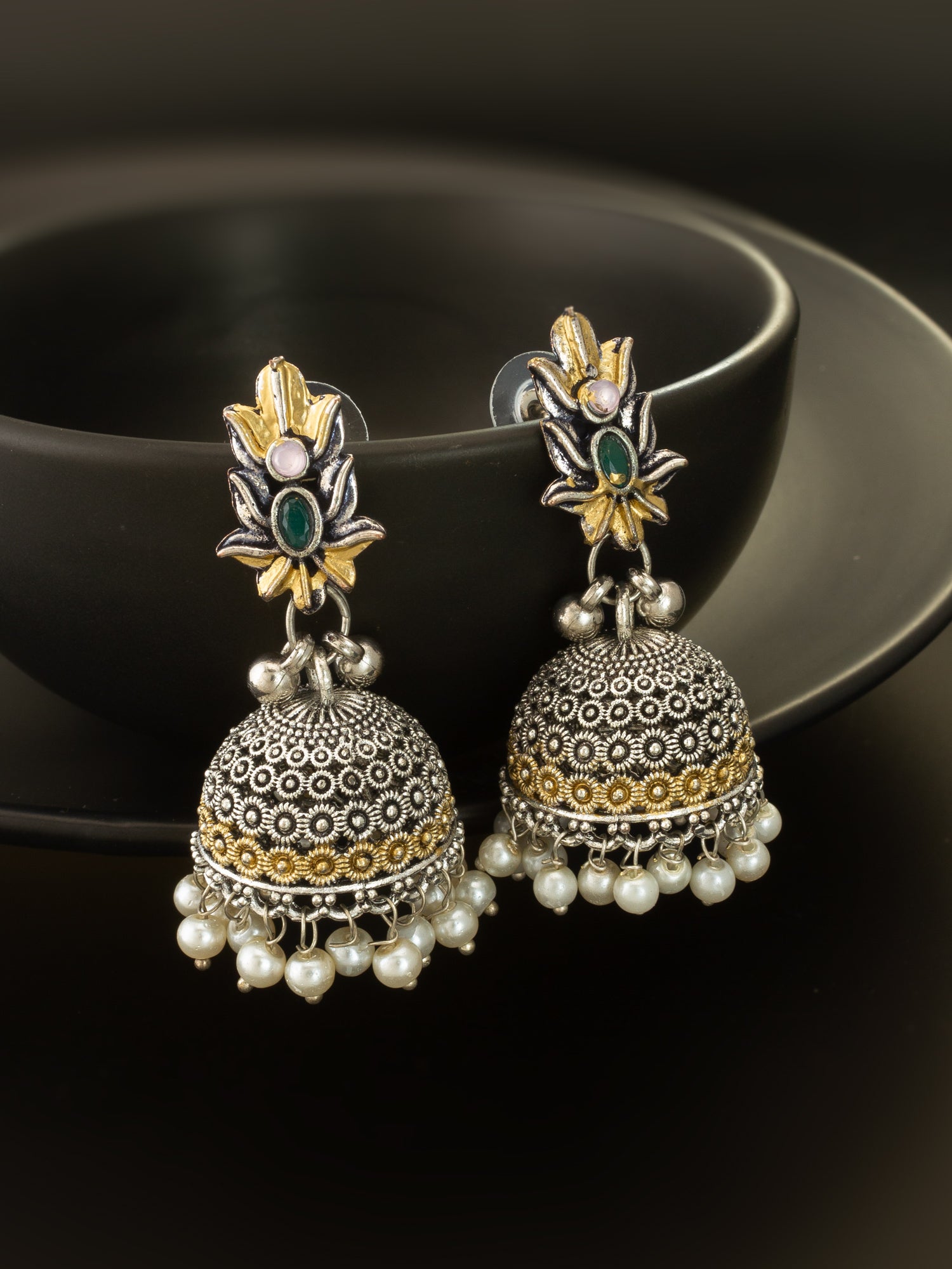 A pair of ornate Indian jhumka earrings with silver filigree bells, gold accents, and small pearl drops, displayed on a black ceramic dish against a dark background.