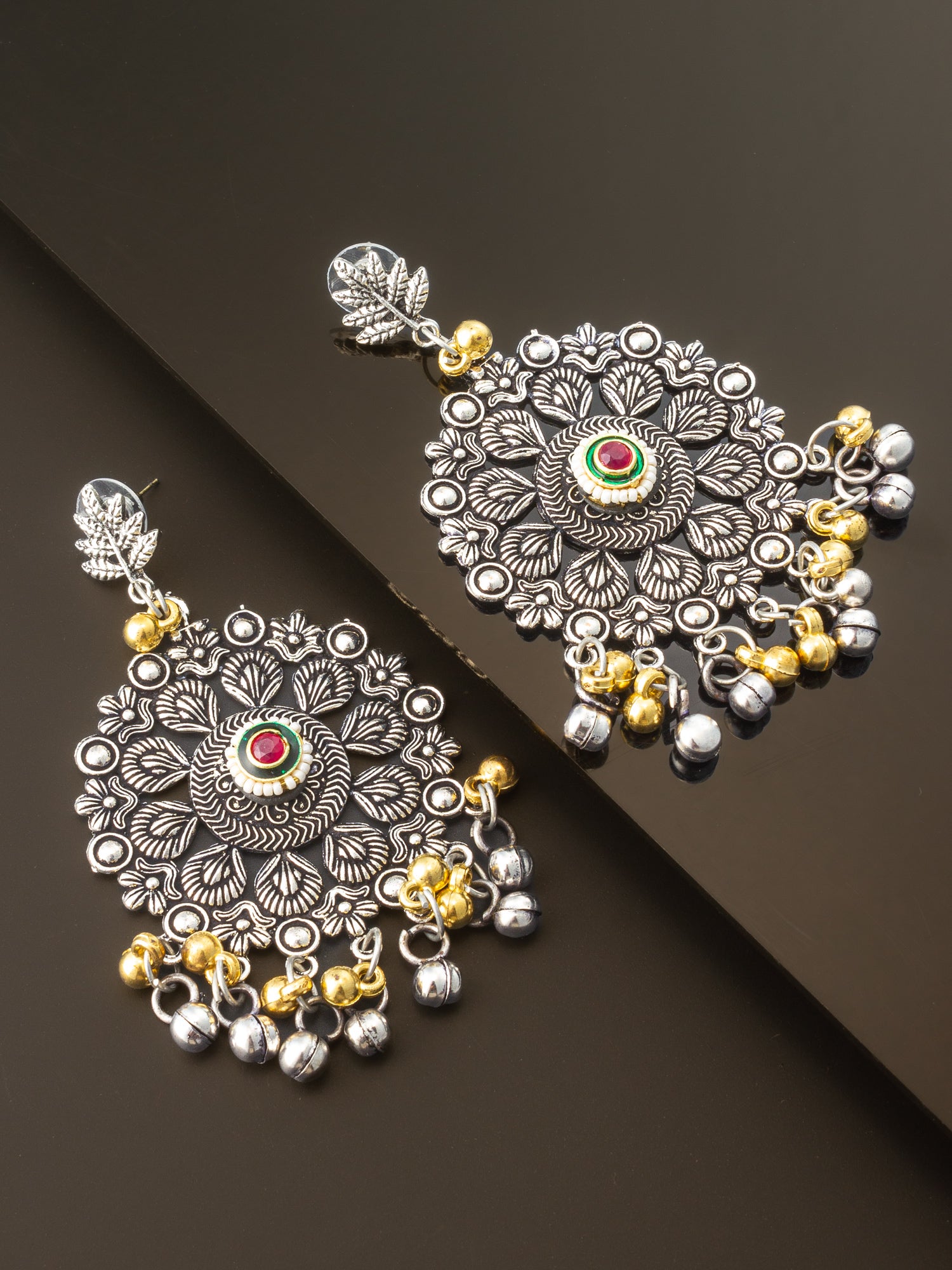 A pair of ornate silver-toned Indian jhumka earrings with gold-toned bell charms, a central red and green stone, and detailed floral engravings, displayed on a dark reflective surface.