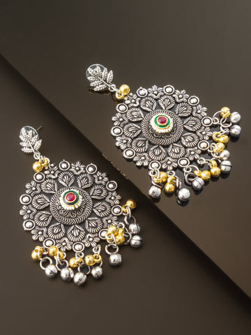 A pair of ornate silver-toned Indian jhumka earrings with gold-toned bell charms, a central red and green stone, and detailed floral engravings, displayed on a dark reflective surface.