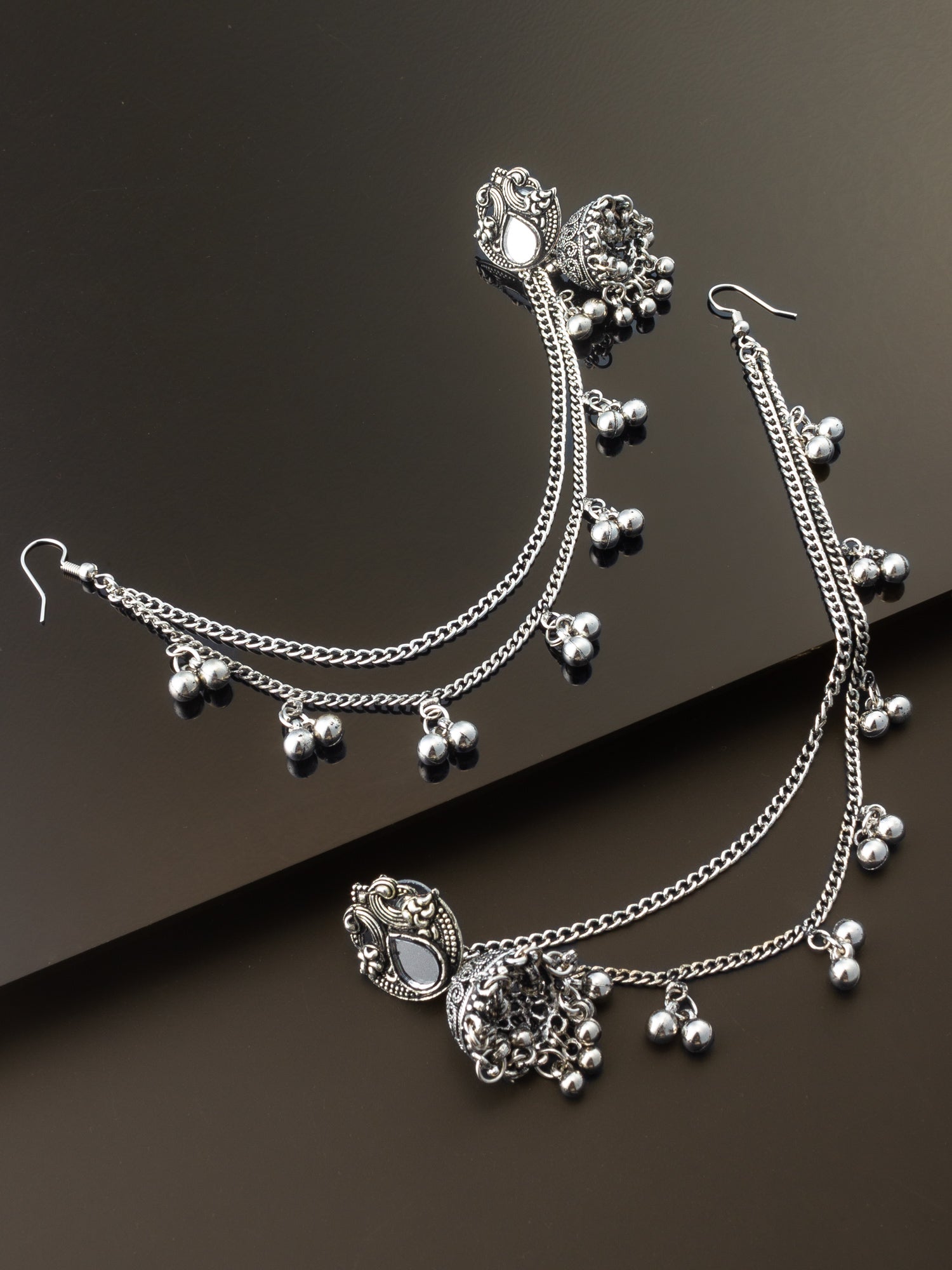 Silver-toned traditional Indian bridal jewelry set featuring a necklace with a detailed pendant, matching dangling earrings, and multiple chain layers adorned with small silver bells, displayed on a dark brown background.