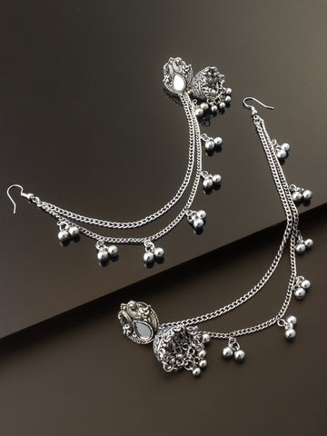 Silver-toned traditional Indian bridal jewelry set featuring a necklace with a detailed pendant, matching dangling earrings, and multiple chain layers adorned with small silver bells, displayed on a dark brown background.