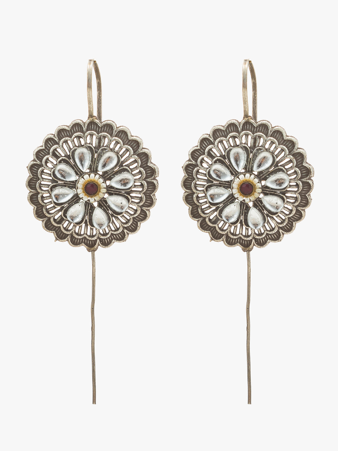 A pair of silver-toned traditional earrings with intricate floral filigree patterns, white inlay stones, and a small red gemstone in the center, displayed on a dark brown background.