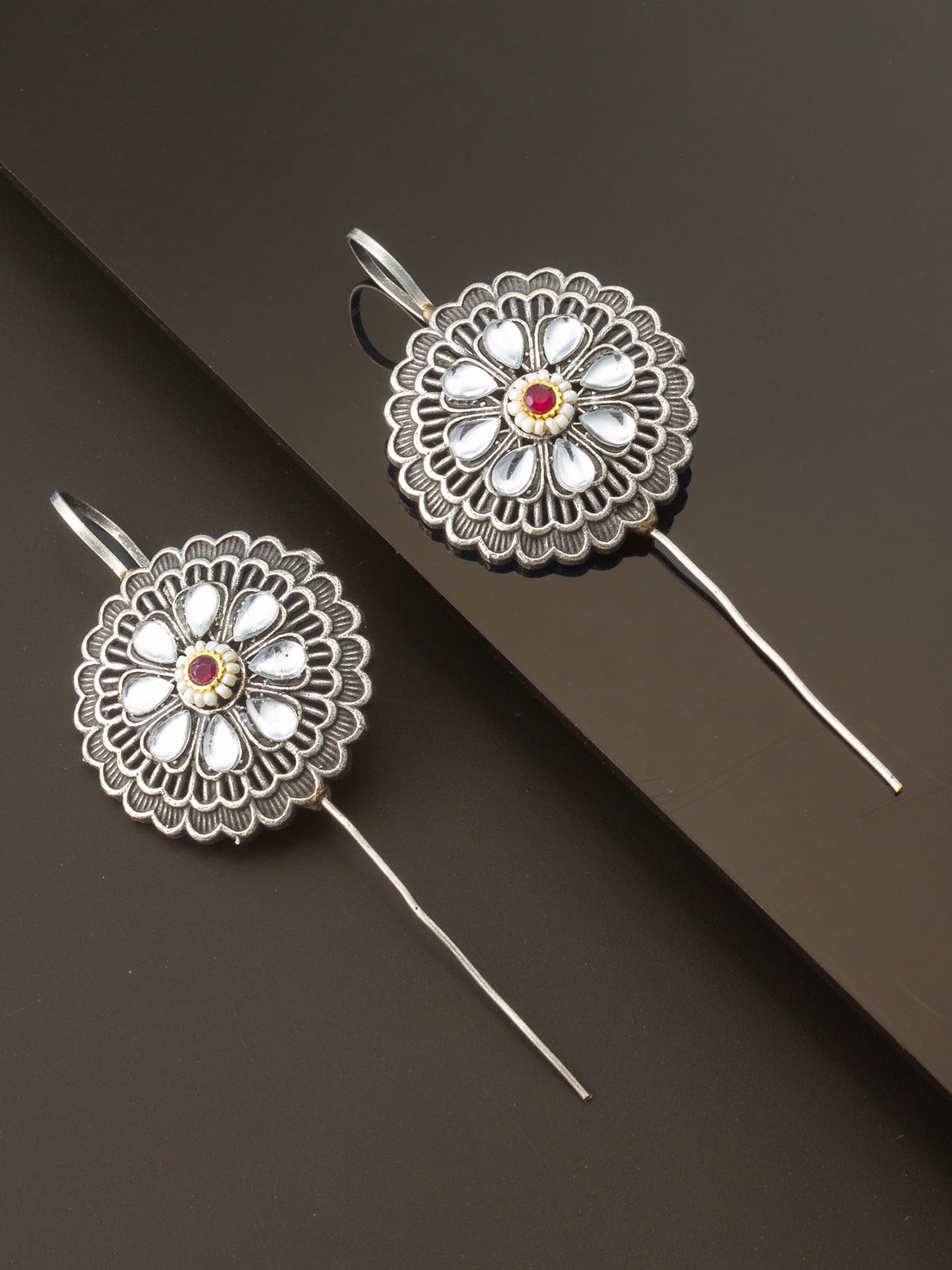 A pair of silver-toned traditional earrings with intricate floral filigree patterns, white inlay stones, and a small red gemstone in the center, displayed on a dark brown background.