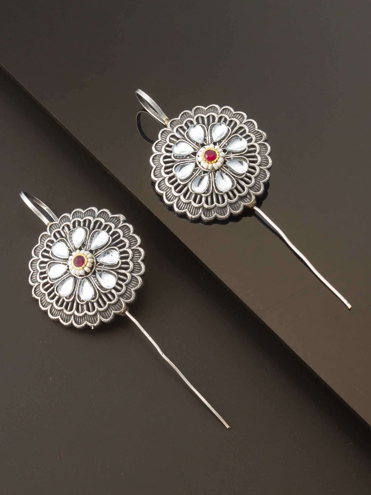 A pair of silver-toned traditional earrings with intricate floral filigree patterns, white inlay stones, and a small red gemstone in the center, displayed on a dark brown background.