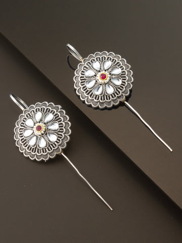 A pair of silver-toned traditional earrings with intricate floral filigree patterns, white inlay stones, and a small red gemstone in the center, displayed on a dark brown background.