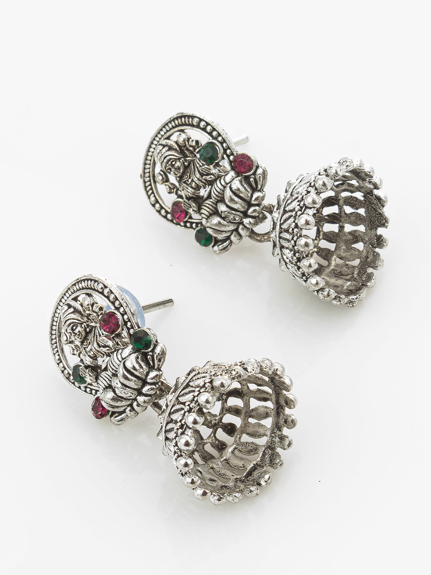 A pair of silver-toned Indian jhumka earrings with detailed filigree work, small green and red gemstone accents, and textured bell-shaped drops on a plain white background.