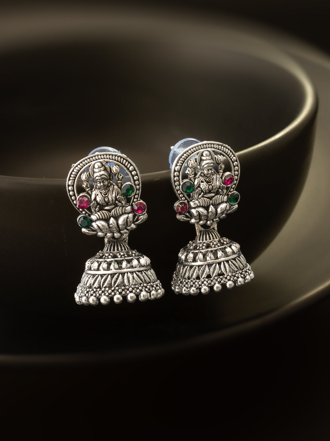 A pair of silver-toned Indian jhumka earrings featuring detailed deity designs and small green and red gemstone accents, displayed on a dark reflective surface.