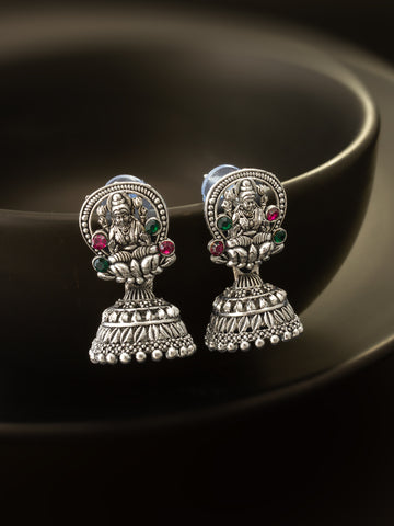 A pair of silver-toned Indian jhumka earrings featuring detailed deity designs and small green and red gemstone accents, displayed on a dark reflective surface.