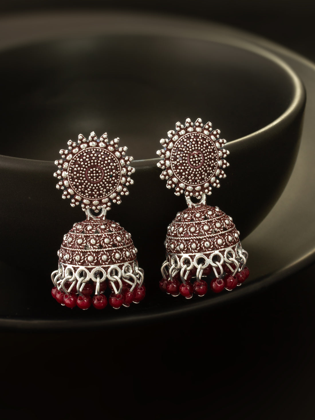 A pair of traditional Indian jhumka earrings with silver-toned metalwork, red stones, and dangling red bead tassels, displayed on a dark glossy surface.