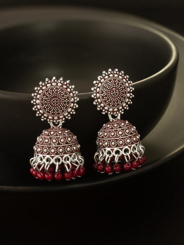A pair of traditional Indian jhumka earrings with silver-toned metalwork, red stones, and dangling red bead tassels, displayed on a dark glossy surface.