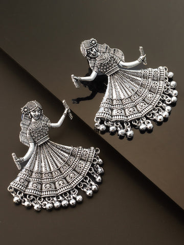 A pair of silver-toned jhumka earrings shaped like dancing figures, displayed on a dark brown background with one earring resting on a reflective surface.