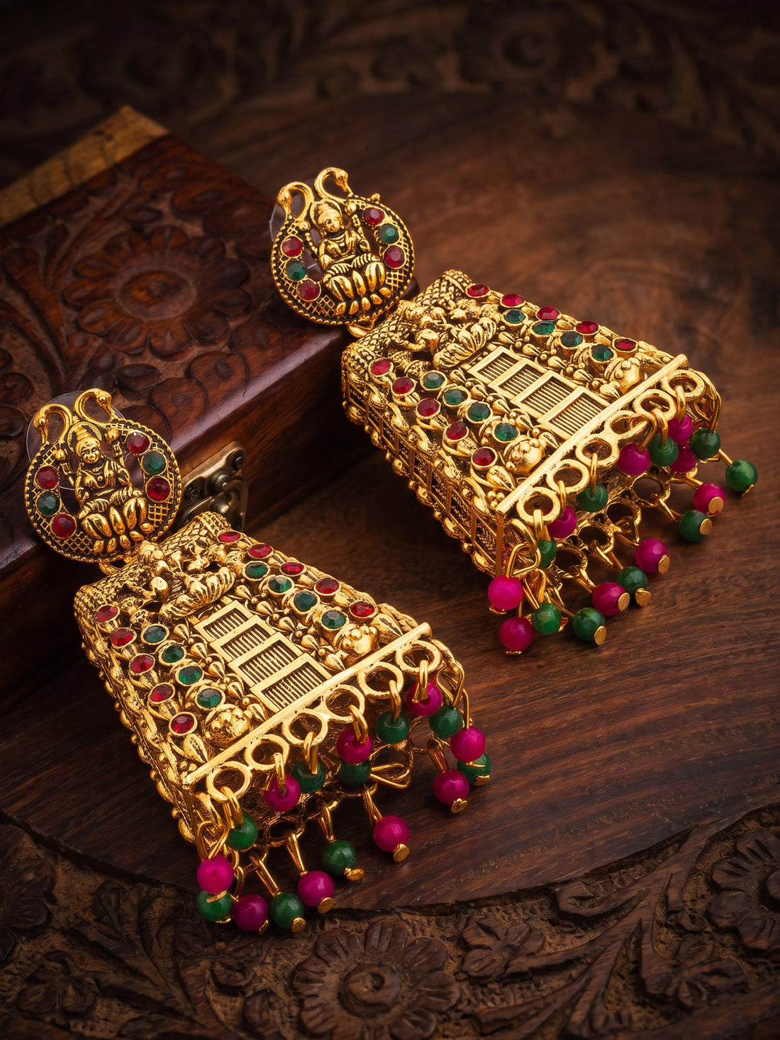 A pair of ornate gold-toned Indian jhumka earrings decorated with red and green stones and dangling beaded tassels, displayed on a carved dark wood surface.