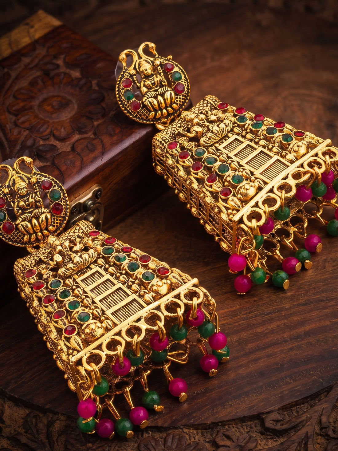 A pair of ornate gold-toned Indian jhumka earrings decorated with red and green stones and dangling beaded tassels, displayed on a carved dark wood surface.