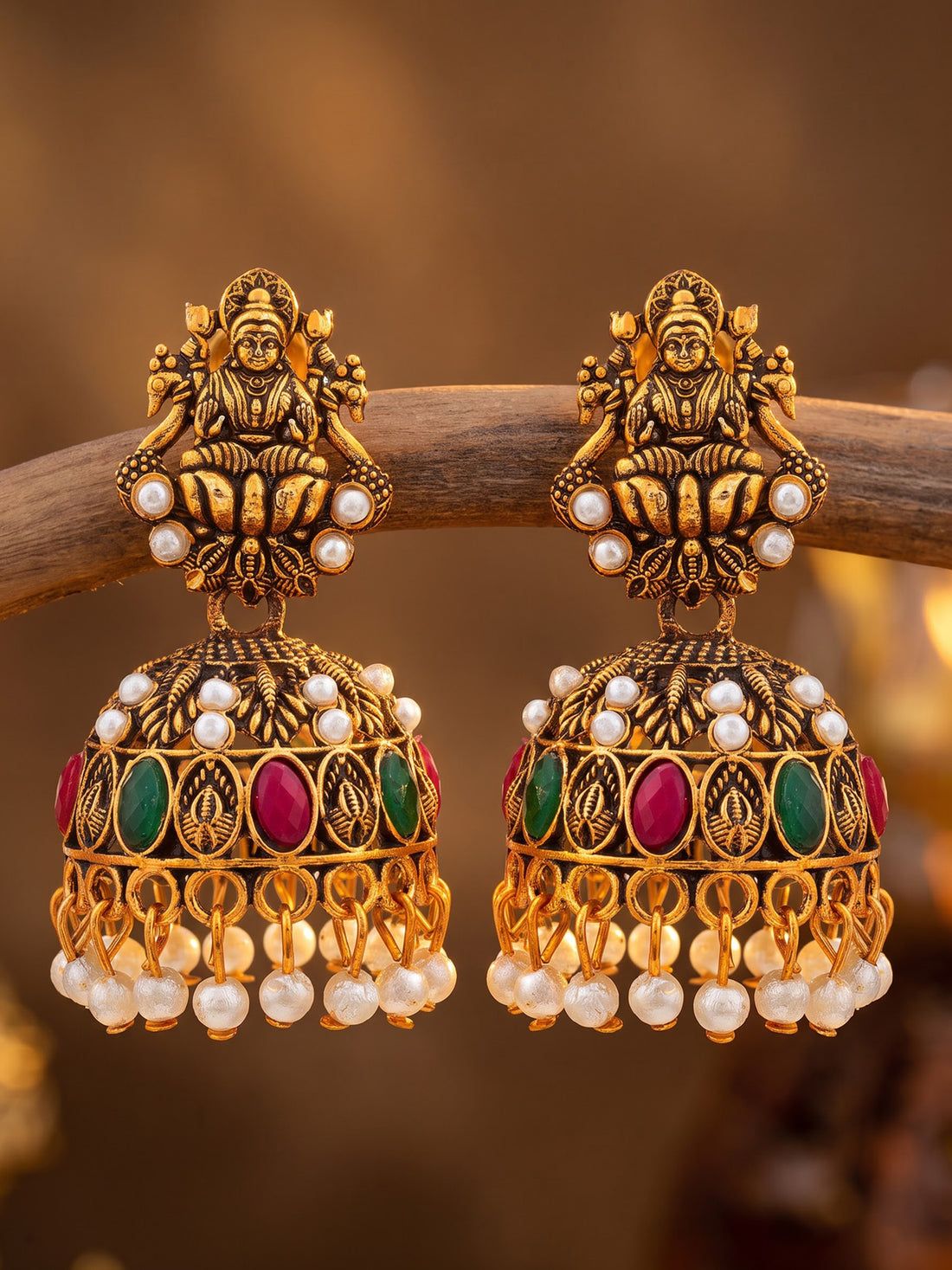 A pair of ornate gold-toned Indian jhumka earrings with deity motifs, white pearl beads, and green and pink gemstone accents, hanging from a wooden branch against a warm brown background.