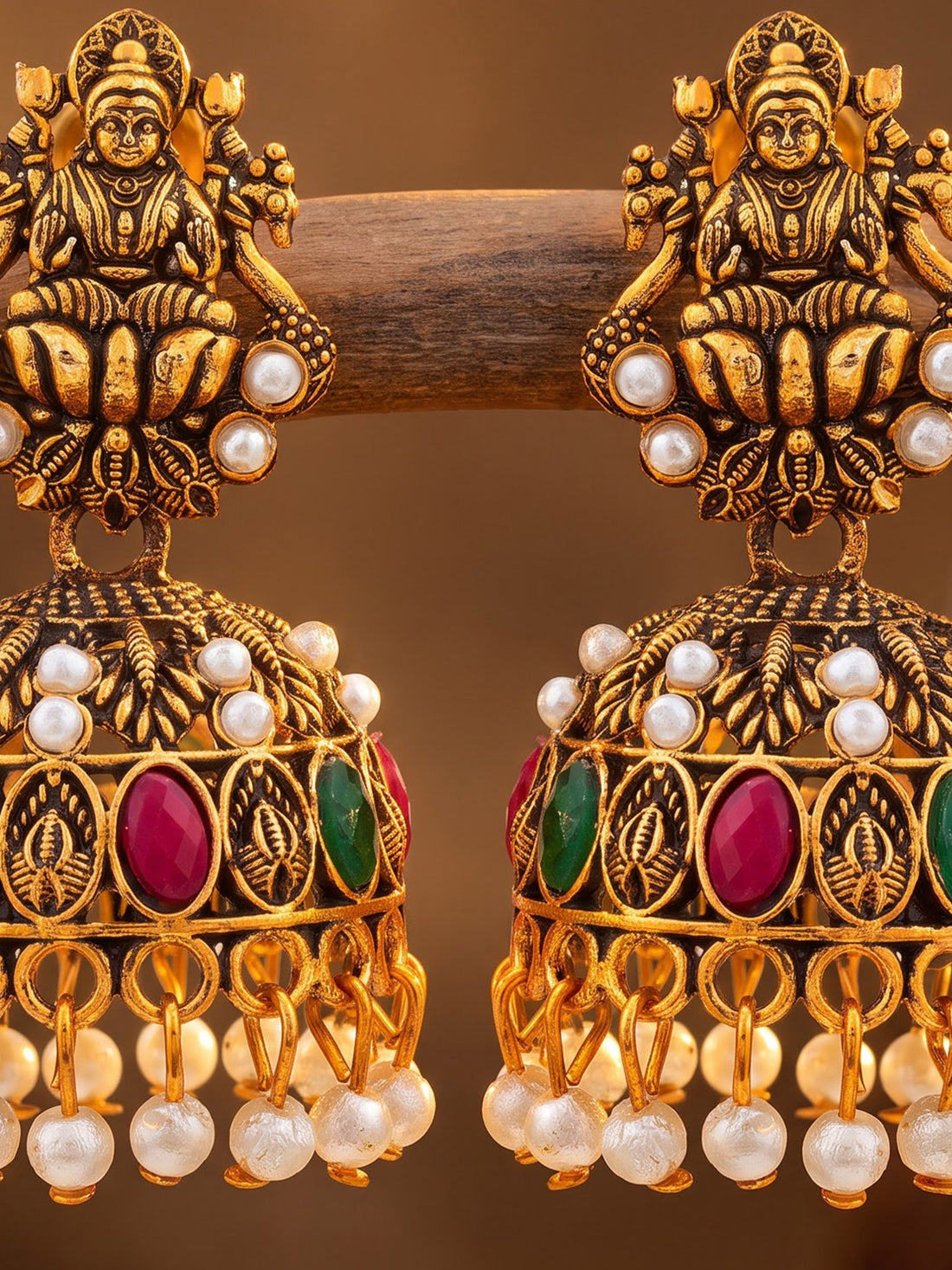 A pair of ornate gold-toned Indian jhumka earrings with deity motifs, white pearl beads, and green and pink gemstone accents, hanging from a wooden branch against a warm brown background.