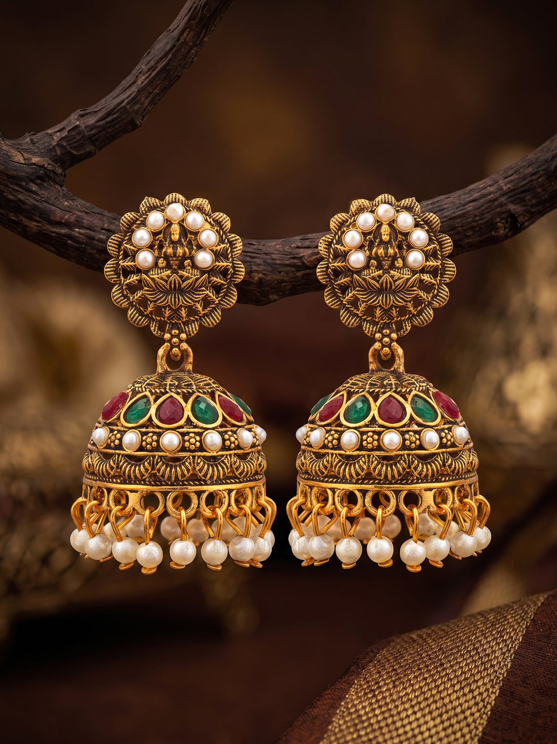 A pair of ornate gold-toned Indian jhumka earrings with white pearl bead borders, green and red enamel accents, and dangling pearl beads, displayed on a dark branch against a warm blurred background.