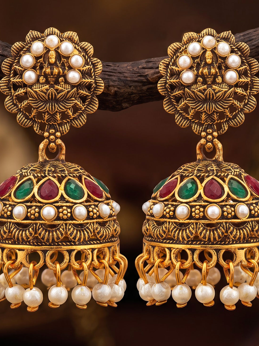 A pair of ornate gold-toned Indian jhumka earrings with white pearl bead borders, green and red enamel accents, and dangling pearl beads, displayed on a dark branch against a warm blurred background.