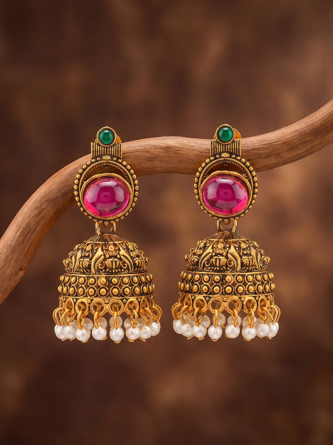 A pair of ornate gold-toned Indian jhumka earrings with large pink stones, small green stones, and dangling white pearl beads, displayed on a curved wooden branch against a warm brown background.