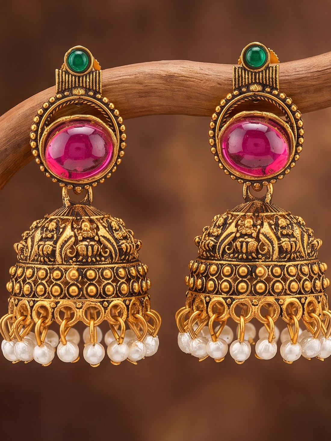 A pair of ornate gold-toned Indian jhumka earrings with large pink stones, small green stones, and dangling white pearl beads, displayed on a curved wooden branch against a warm brown background.