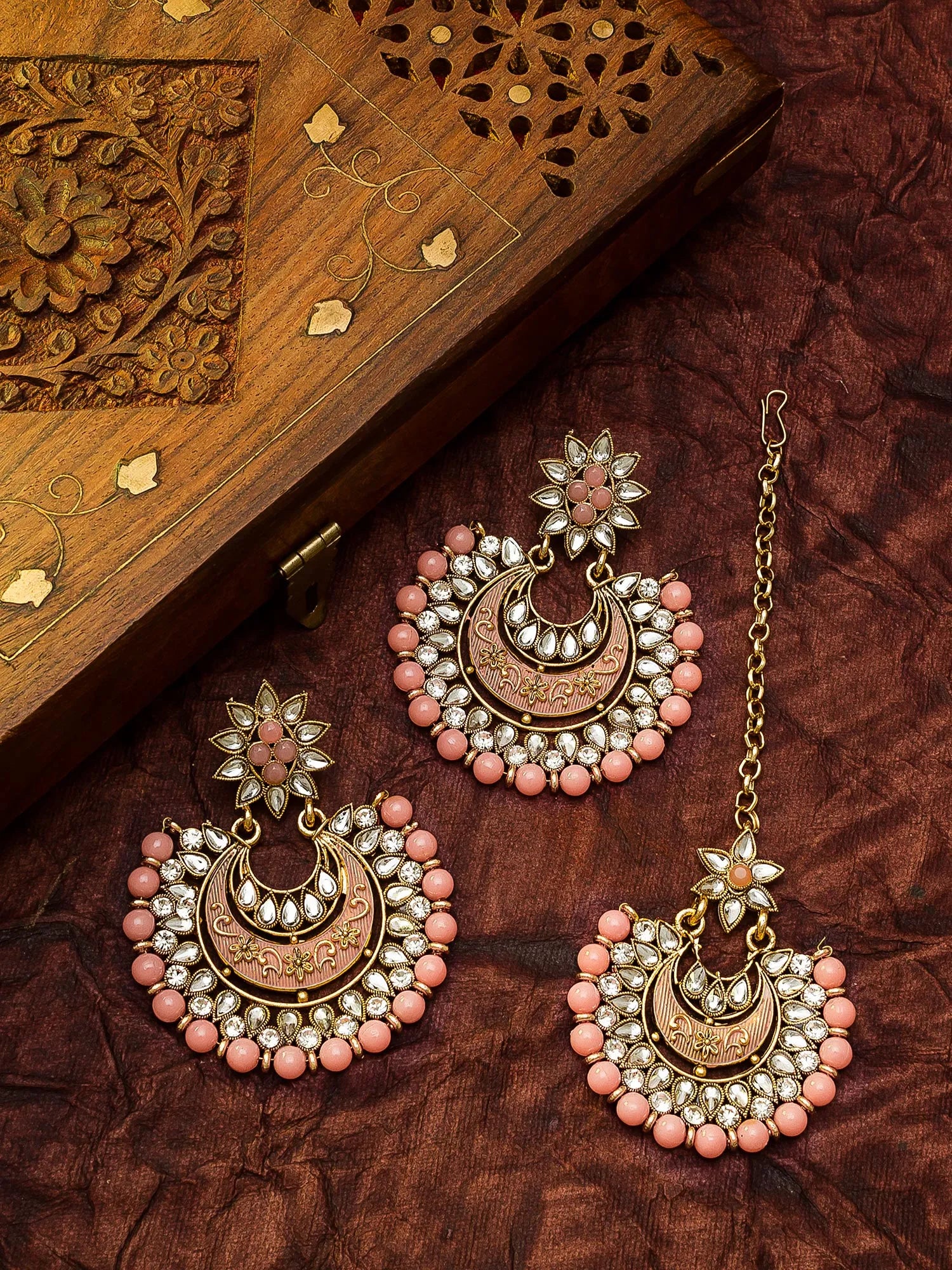 Gold-toned traditional Indian jhumka earrings with pink stone accents and white stones, displayed alongside a matching chain and a carved wooden box on a textured dark brown fabric background.