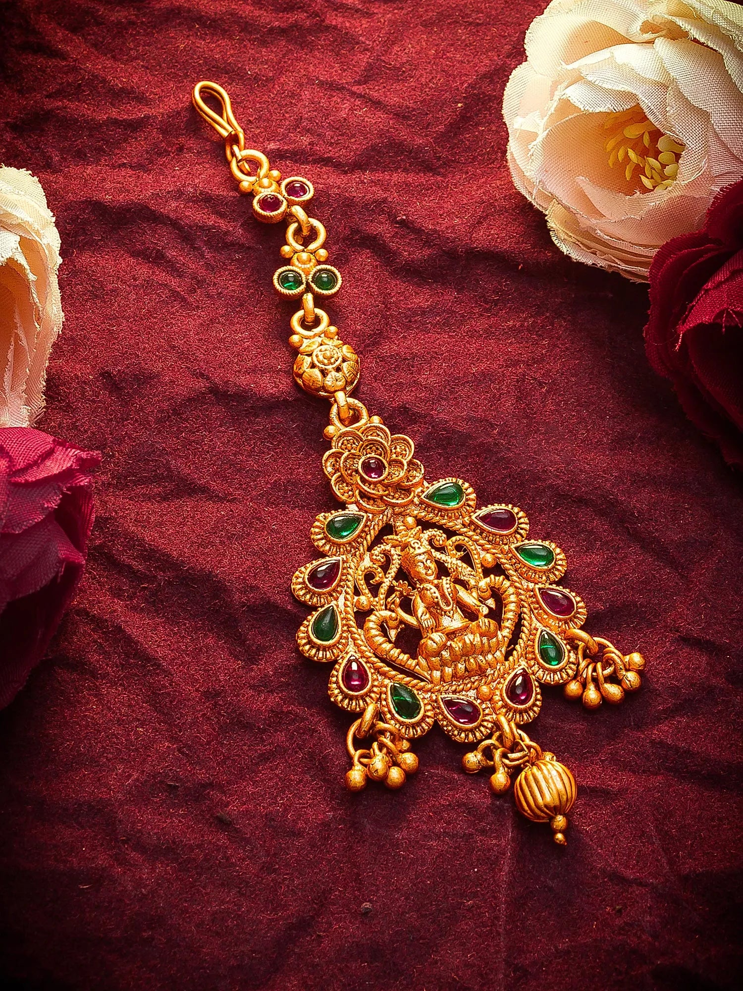 An ornate gold-toned traditional Indian jewelry piece, likely a maang tikka or pendant, decorated with green and red stones, displayed on a textured dark red fabric background with decorative flowers.