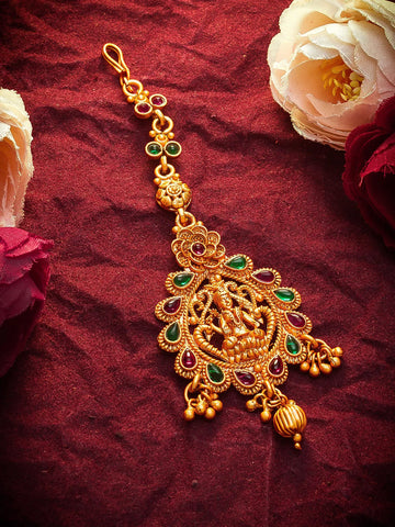 An ornate gold-toned traditional Indian jewelry piece, likely a maang tikka or pendant, decorated with green and red stones, displayed on a textured dark red fabric background with decorative flowers.