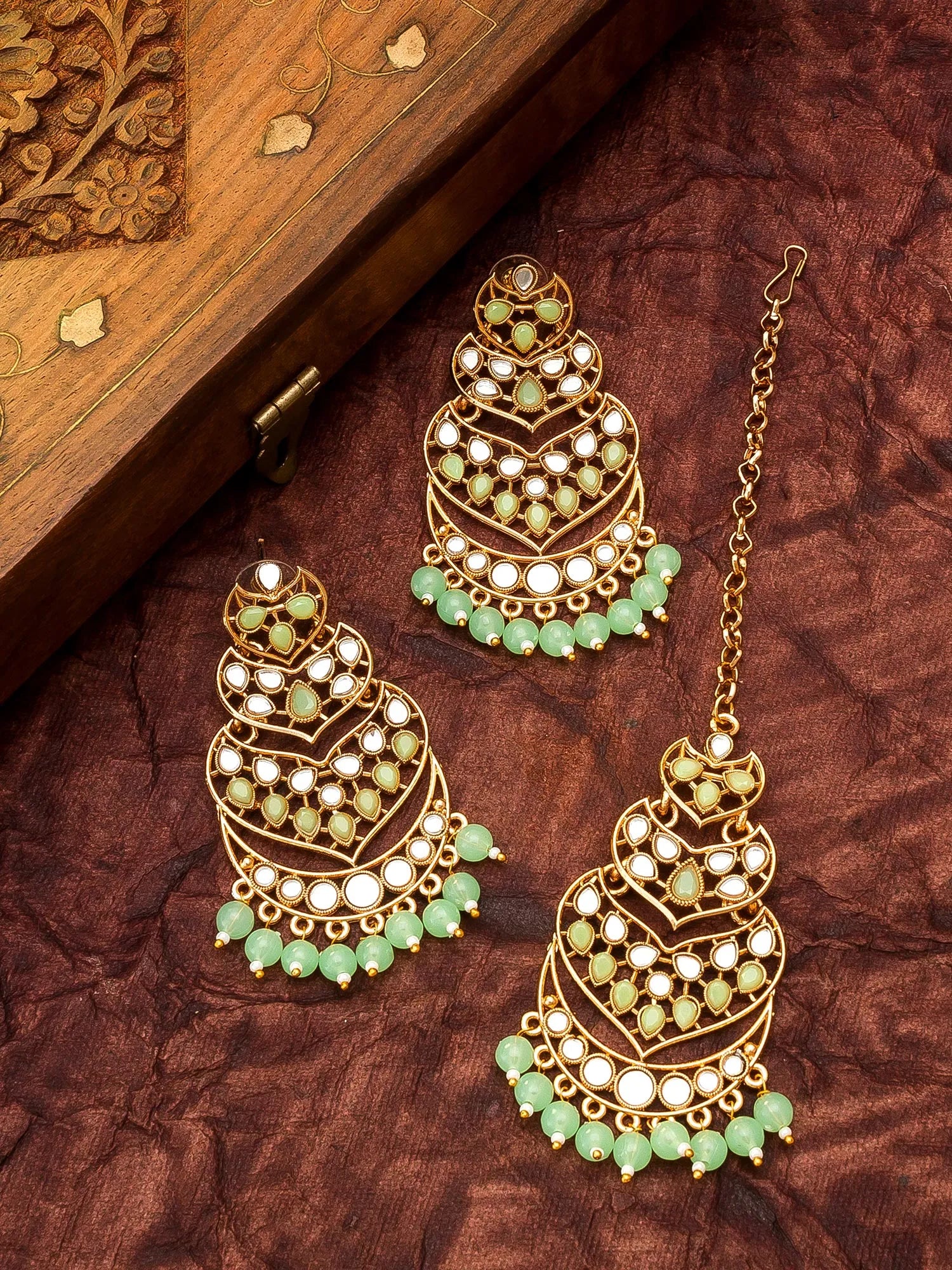 Gold-toned traditional Indian jhumka earrings with green stone accents and white pearl-like details, displayed on a textured brown fabric next to a carved wooden box.