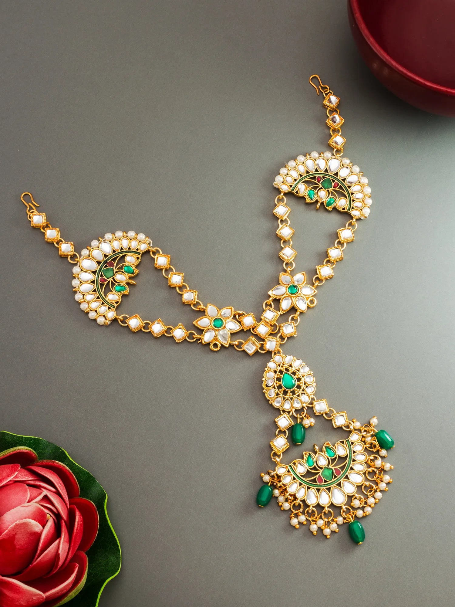 Gold-plated Indian traditional jewelry set featuring a maang tikka and matching chain with white faux pearls, green stone accents, and dangling green bead details, displayed on a gray background with decorative red rose and bowl props.