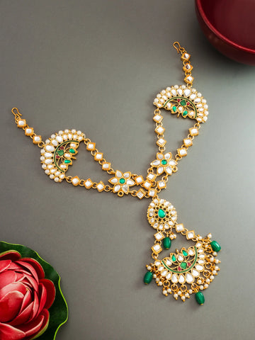 Gold-plated Indian traditional jewelry set featuring a maang tikka and matching chain with white faux pearls, green stone accents, and dangling green bead details, displayed on a gray background with decorative red rose and bowl props.