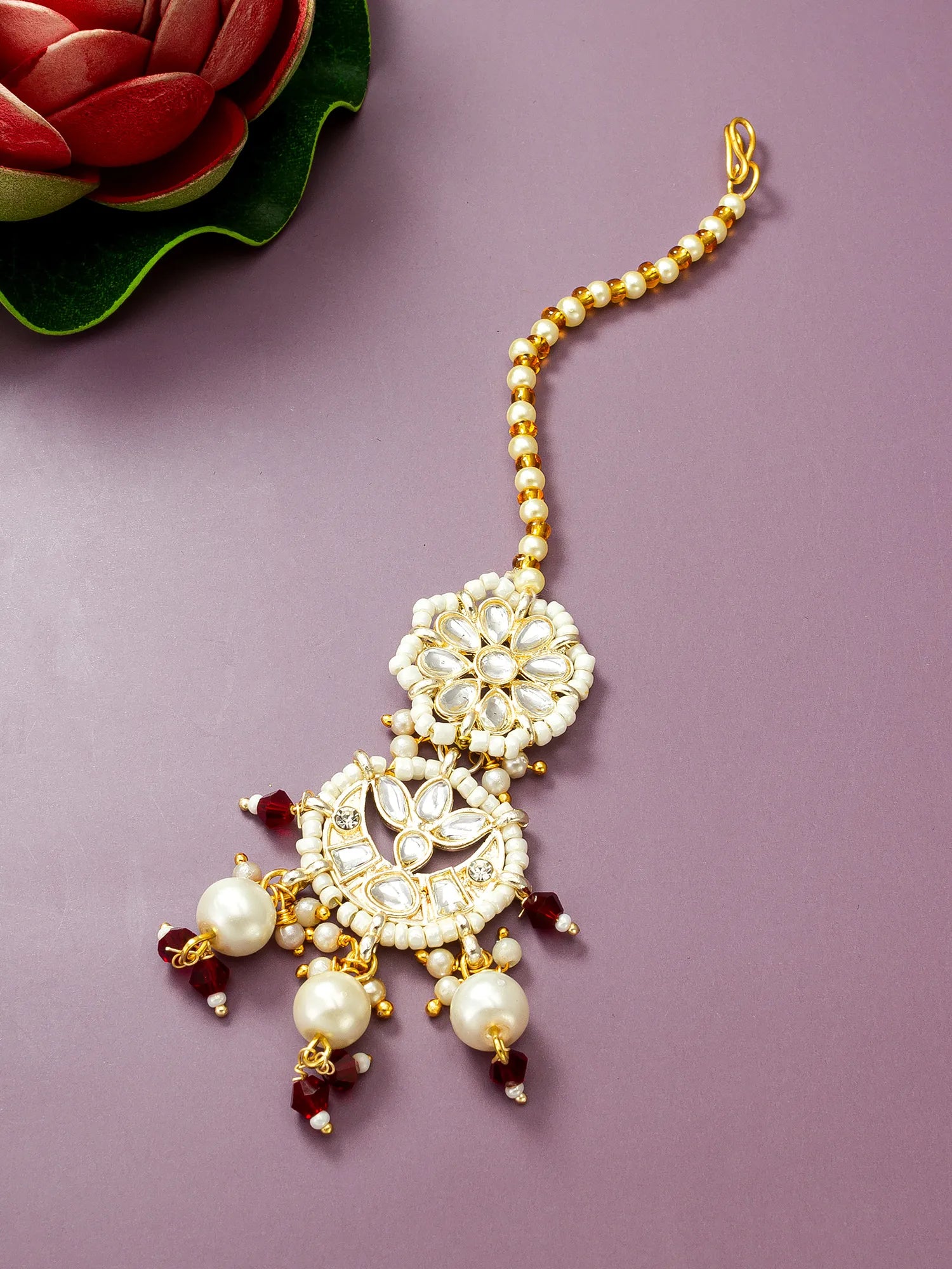 Gold and white pearl Indian maang tikka with hanging maroon bead accents, displayed on a dusty purple background with a decorative red lotus leaf in the corner.