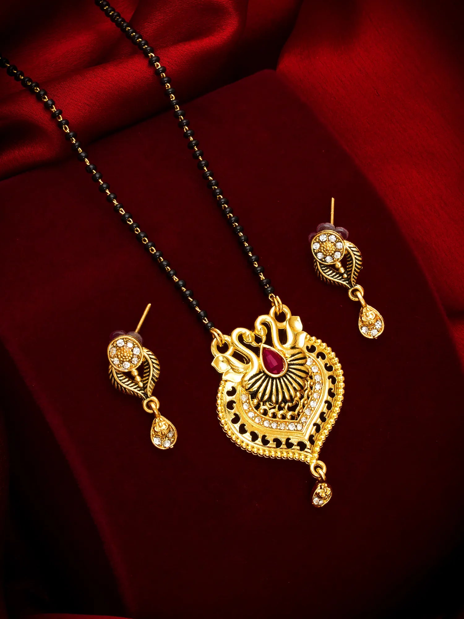 Gold-toned traditional Indian jewelry set featuring a necklace with a detailed heart-shaped pendant, a black beaded chain, and matching drop earrings, displayed on a rich red satin background.