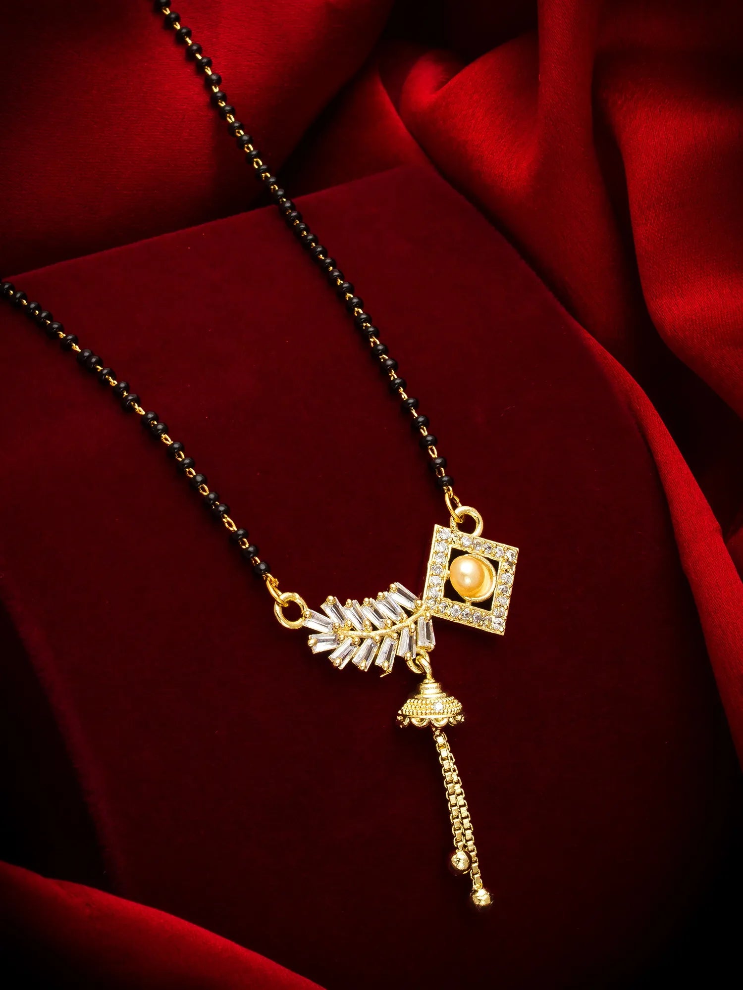A gold and black beaded necklace with a decorative pendant featuring a square crystal frame, a gold-toned bell charm, and a feather-like element, displayed on a rich red satin fabric background.